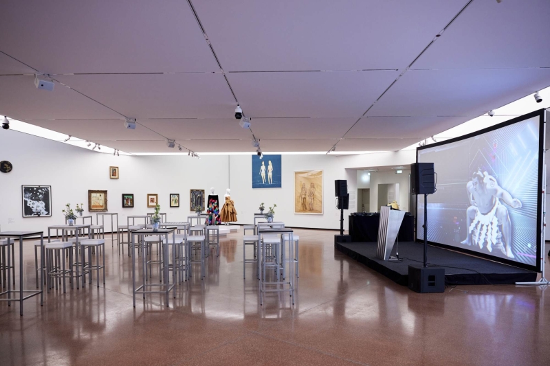 Modern art gallery with tables, artworks on walls, and a large screen displaying animated visuals.