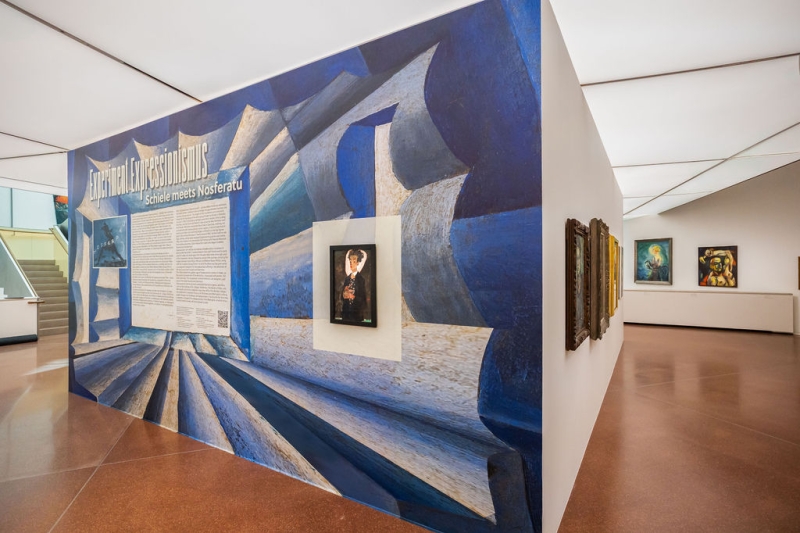 The photo shows the exhibition “Experiment Expressionism. Schiele meets Nosferatu” at the Heidi Horten Collection.
At the front, there is a big wall in blue and white.
On the wall, there is large writing: “Experiment Expressionism – Schiele meets Nosferatu”.
In the center, there is a small painting by Egon Schiele – a self-portrait.
Next to it, there is a small picture from the old film “Nosferatu”.
Below, there is a long text about the exhibition.
Behind the wall, you can see more colorful paintings on white walls.
The room is bright and open.
The ceiling is white, and the floor is brown.
The exhibition looks modern, clear, and well organized.