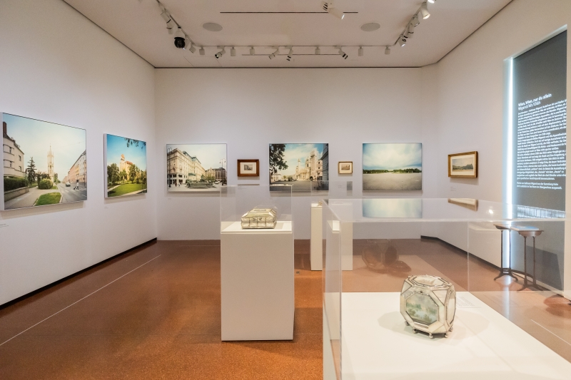 The photo shows an exhibition room with many pictures on the walls and three display cases in the middle. On display is a cross-section of historical and contemporary views of Vienna.

Large-format photographs by Stefan Oláh hang on the walls, showing Viennese squares, streets and buildings in a calm, clear visual language. In between are watercolors by Rudolf von Alt.

In the display cases are tins with views of Vienna by Balthasar Wigand.  Each box shows several views - of St. Stephen's Cathedral or the Danube, for example.

There is an exhibition text on the right-hand wall.
