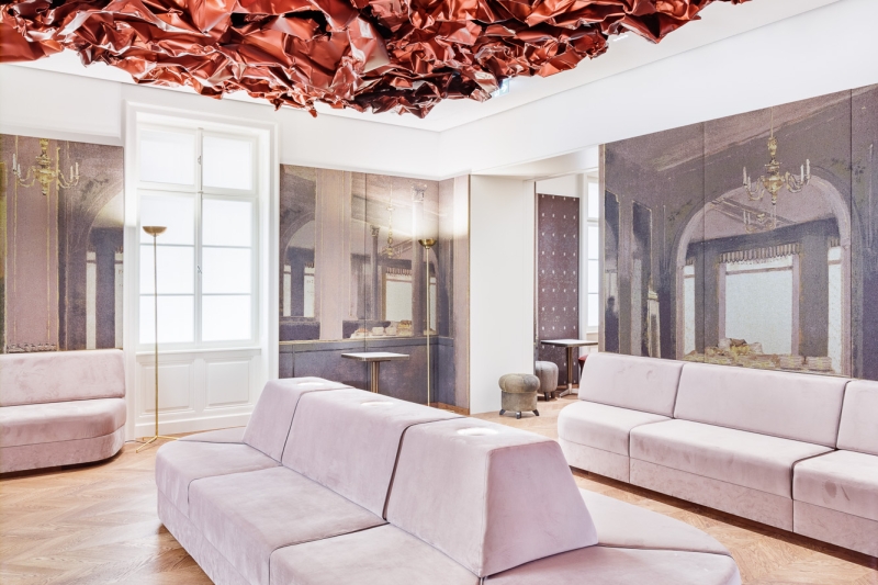 Auf dem Foto sehen wir den Tea Room im Museum der Heidi Horten Collection in Wien.

An der Decke hängt ein großes, rotes Kunstwerk von Hans Kupelwieser.
Es sieht aus wie zerknittertes Papier oder glänzender Stoff,
wurde aber aus Aluminium gemacht.

An der hinteren Wand gibt es viele runde Fenster – sie sehen aus wie Bullaugen.
Dahinter befinden sind kleine, kostbare Objekte aus der Sammlung von Heidi Horten.

Diese Vitrinenwand wurde von Markus Schinwald entworfen.
Auch die rosa Sitzmöbel in der Mitte des Raums gehören zu seinem Konzept.
Sie laden ein, sich hinzusetzen, zu schauen und zu entspannen.

Die Wände sind mit großen Stoffbildern bedeckt,
die an ein altes Café oder Schlosszimmer erinnern.