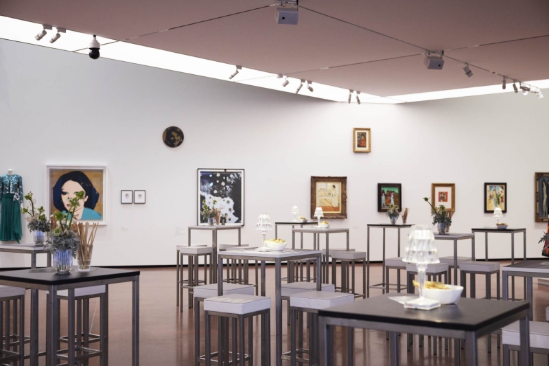 Art gallery interior featuring various framed artworks, modern tables with floral arrangements, and a green dress display.