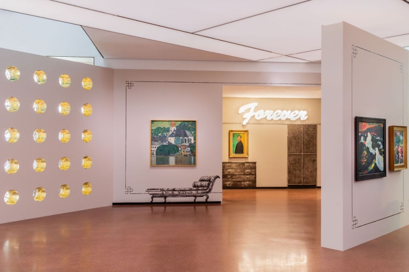 The photo shows part of the “Klimt⇄Warhol” exhibition in the Heidi Horten Collection museum in Vienna.

On the left you can see a wall with round windows that look like portholes.
This wall was designed by the artist Markus Schinwald.

In the middle, directly in front of us, hangs a painting by Gustav Klimt.
It shows the Attersee, a lake in Austria.
The picture shows water, houses and nature in bright colors.

Markus Schinwald also designed the decorations on the walls and the painted pieces of furniture next to and under the works of art.

At the back of the wall, above a picture, the word “Forever” is illuminated in large white letters.
This illuminated lettering is a work of art by Tim Noble & Sue Webster.
Below it hangs a self-portrait by Edvard Munch.

On the far right, another wall juts diagonally into the room.
Two paintings by Emil Nolde, which are also part of the collection, hang there.