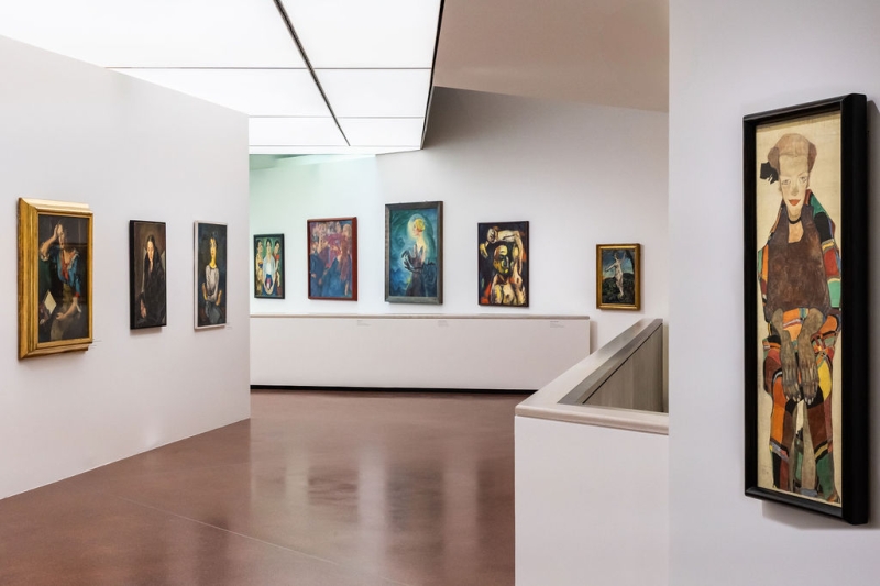 The photo shows a bright room in the exhibition “Experiment Expressionism.”
There are many paintings hanging on the white walls.
The pictures show women and men – some look calm, others are moving or appear strong.

On the far right, you see a long, narrow painting of a woman.
The painting is by Egon Schiele.

On the left wall, there are three portraits hanging next to each other.
In the middle of the room, you see more paintings, for example the picture “Terzetta”, with the woman and the long hands.
The floor is brown, and the ceiling is bright.