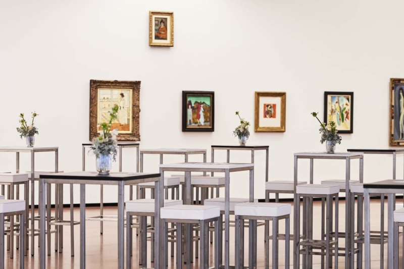 Gallery space with tall tables, white stools, and various framed artworks on the walls, featuring floral centerpieces.