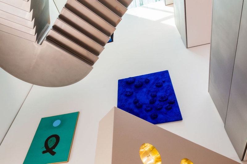 The photo shows a view upstairs in the Heidi Horten Collection museum in Vienna.
You can see a modern metal staircase and large, bright walls.

On the left wall hangs a green picture with a black sign and a blue circle.
This work of art is called “Blue Orb” and is by Adolph Gottlieb.

Next to it, further up, you can see a bright blue work of art with sponges on it.
This is the very first sponge relief by Yves Klein.
He has dyed real sponges with deep blue paint and glued them onto the canvas.
The work is very special because Yves Klein became famous for this intense blue color.

At the bottom of the picture you can see part of Markus Schinwald's porthole wall.
The round, shiny gold windows are part of a design element.