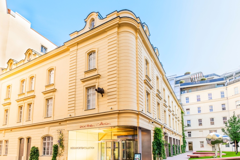 Elegant yellow building with large glass entrance, greenery, and modern sculptures in a sunny courtyard.