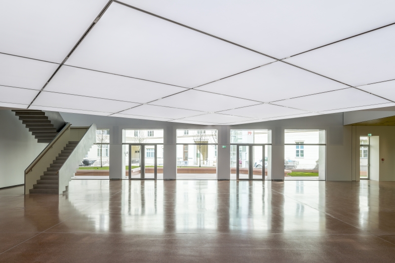 Spacious interior with a glossy floor, large windows, and a staircase on the left, under a bright, grid-patterned ceiling.