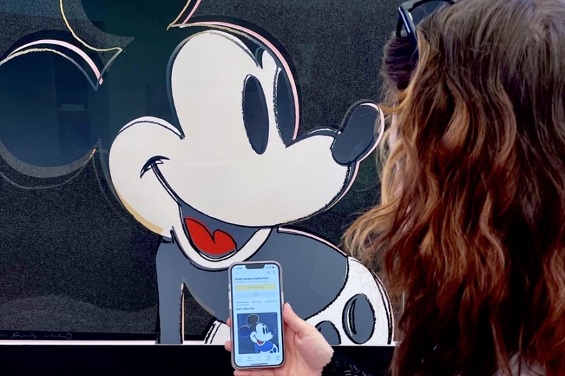 Auf dem Foto sieht man eine Frau von hinten, die sich ein Kunstwerk von Andy Warhol anschaut. Auf dem Bild ist Micky Maus. Die Frau h&auml;lt ein Smartphone in der Hand. Darauf ist die App Smartify ge&ouml;ffnet &ndash; ein Audioguide, der Informationen &uuml;ber Kunstwerke gibt. Man sieht, dass sie genau das Bild auf dem Handy ge&ouml;ffnet hat, das auch vor ihr an der Wand h&auml;ngt. Das Foto zeigt, wie digitale Medien im Museum helfen, mehr &uuml;ber Kunst zu erfahren &ndash; einfach und direkt am eigenen Handy