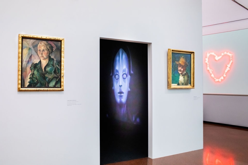 The photo shows a doorway in the exhibition “Experiment Expressionism. Schiele meets Nosferatu”.
In the room behind the doorway, there is a large glowing image of the machine figure from the film “Metropolis” (1927).

On the left and right sides of the doorway, there are two portraits in golden frames:
On the left is a sharp-edged, serious portrait of a woman.
On the right is a smaller, more loosely painted portrait with a blue-green background and a yellow hat – this is the self-portrait of Herbert Boeckl.

In the background on the right, a pink neon heart is glowing on the wall.