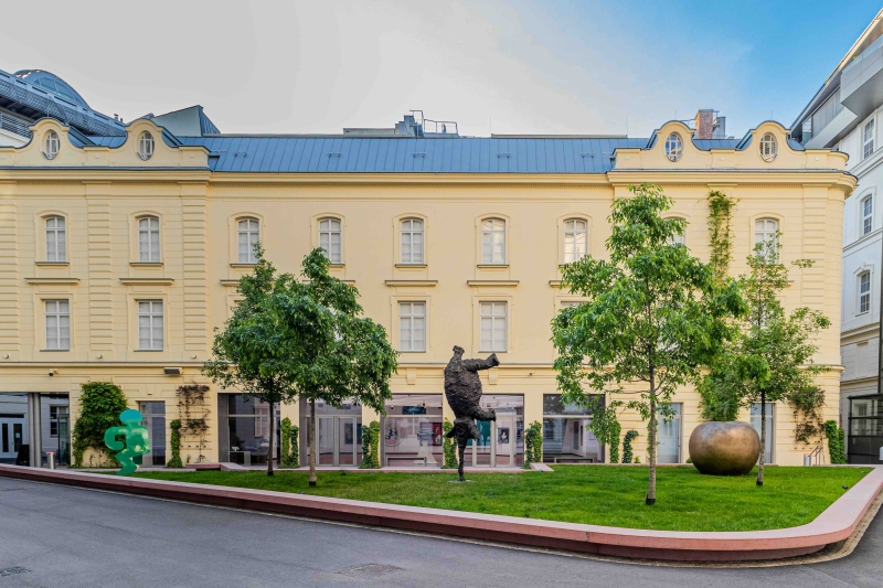 Auf dem Foto sieht man das Gebäude der Heidi Horten Collection in Wien.
Es ist ein hellgelbes Haus mit neoklassizistischer Fassade und großen Fenstern.
Das Gebäude steht im Hanuschhof, einem ruhigen Innenhof mitten in der Stadt.
Vor dem Museum ist ein kleiner Skulpturengarten mit Gras, Bäumen und Kunstwerken.
Man sieht dort drei verschiedene Skulpturen:
Ganz links steht eine grüne Figur von Keith Haring.
In der Mitte steht eine dunkle Bronzefigur, ein Elefant von Miquel Barceló.
Ganz rechts sieht man ein großer goldener Apfel. Er stammt von Claude Lalanne.