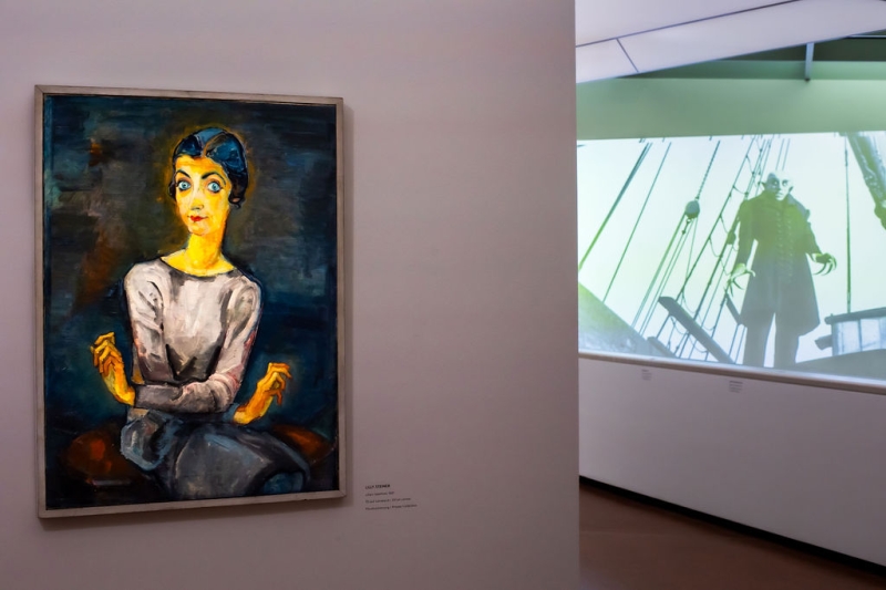 The photo shows a part of the exhibition “Experiment Expressionism.”

On the left, there is a painting by Lilly Steiner.
It shows Lilian Gärtner with big blue eyes and a grey dress.
She is sitting and holding her hands in a special, expressive way.
This painting was already described earlier.

On the right, you see a film scene from “Nosferatu.”
The film is black and white and is projected onto the wall.
You can see the vampire Nosferatu standing on a ship.
He is on the deck and looks scary.