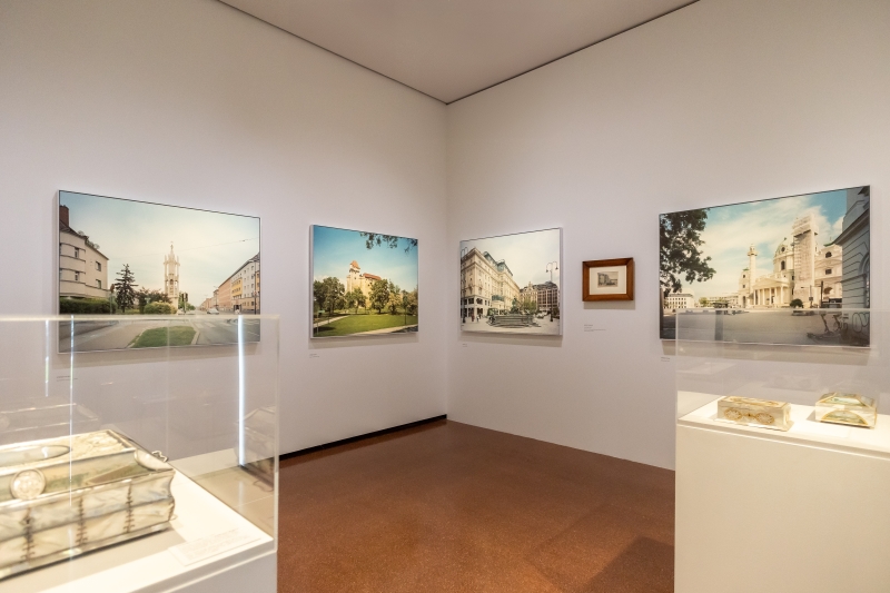 The photo shows an exhibition room with several pictures on the walls and two display cases in the middle. On display is a cross-section of historical and contemporary views of Vienna.

Large-format photographs by Stefan Oláh hang on the walls, showing Viennese squares, streets and buildings in a calm, clear visual language. In between is a watercolor by Rudolf von Alt.

In the display cases are tins with views of Vienna by Balthasar Wigand. Each box shows several views - of St. Stephen's Cathedral or the Danube, for example.