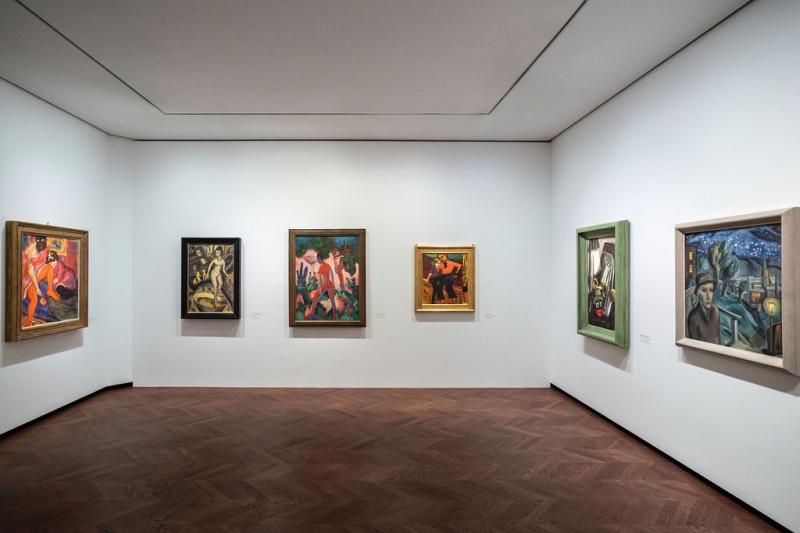 The photo shows a room from the “Experiment Expressionism” exhibition.
The room is bright, with white-painted walls and a brown wooden floor.

Six paintings hang on the walls.
The paintings show people in motion, naked bodies, strong colors and special poses - typical of Expressionism.