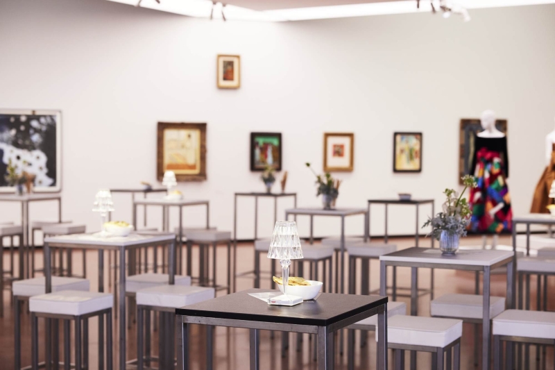 Art gallery with small tables, decorative lamps, and framed artworks on the walls, featuring a colorful dress on display.