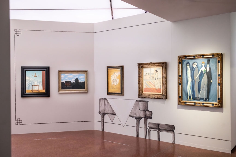 The photo shows part of the "Klimt⇄Warhol" exhibition in the Heidi Horten Collection museum in Vienna.

You can see a wall with several paintings. On the left are two surrealist paintings by René Magritte. 
Below the paintings in the middle is an ornament in the shape of a grand piano. The decorations on the walls and the painted pieces of furniture were designed by the artist Markus Schinwald.