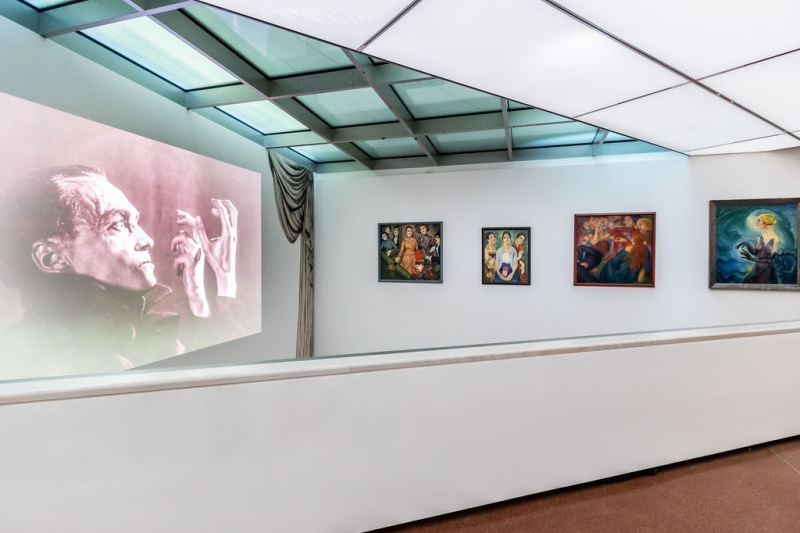 On the far left, you see a photo projected onto a wall.
It shows the actor Conrad Veidt in black and white.
He is raising his hands and looks serious.

To the right of it, there are four paintings:

– The first one on the left shows several people. In the center, a woman sits in an orange dress.
– The second one shows three women.
– The third one shows people from the story Decameron.
– The fourth is the painting “Terzetta”, with the woman raising her long, dark hands.

The colors in all the paintings are strong and bright.