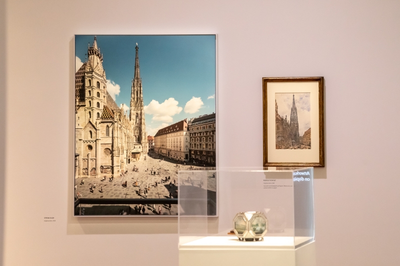 The photo shows an exhibition view with three works of art that deal with the city of Vienna.

On the left is a large photograph by Stefan Olah. It shows St. Stephen's Cathedral in fine weather - with a blue sky and a view of the busy square in front of it.

To the right hangs a smaller watercolor by Rudolf von Alt. This also shows St. Stephen's Cathedral - finely painted, with warm colors and many details.

In the display case below is a Wigand box. It shows various views of Vienna, including St. Stephen's Cathedral.
