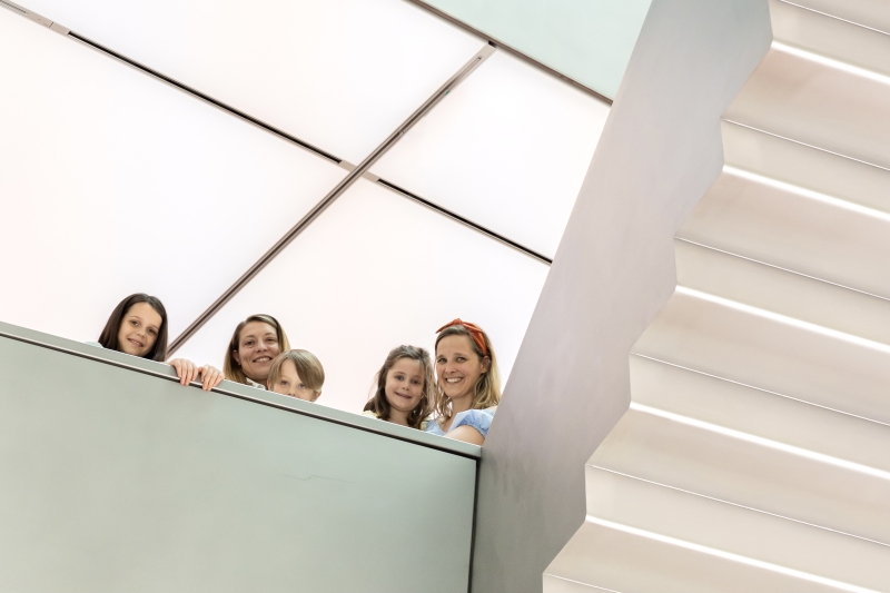 The photo shows five people, including three children and two adults. They are standing on an exhibition level and looking at the camera in a friendly manner. They are all smiling.

On the right of the picture you can see a modern, bright staircase. Behind the people you can see the white illuminated ceiling.