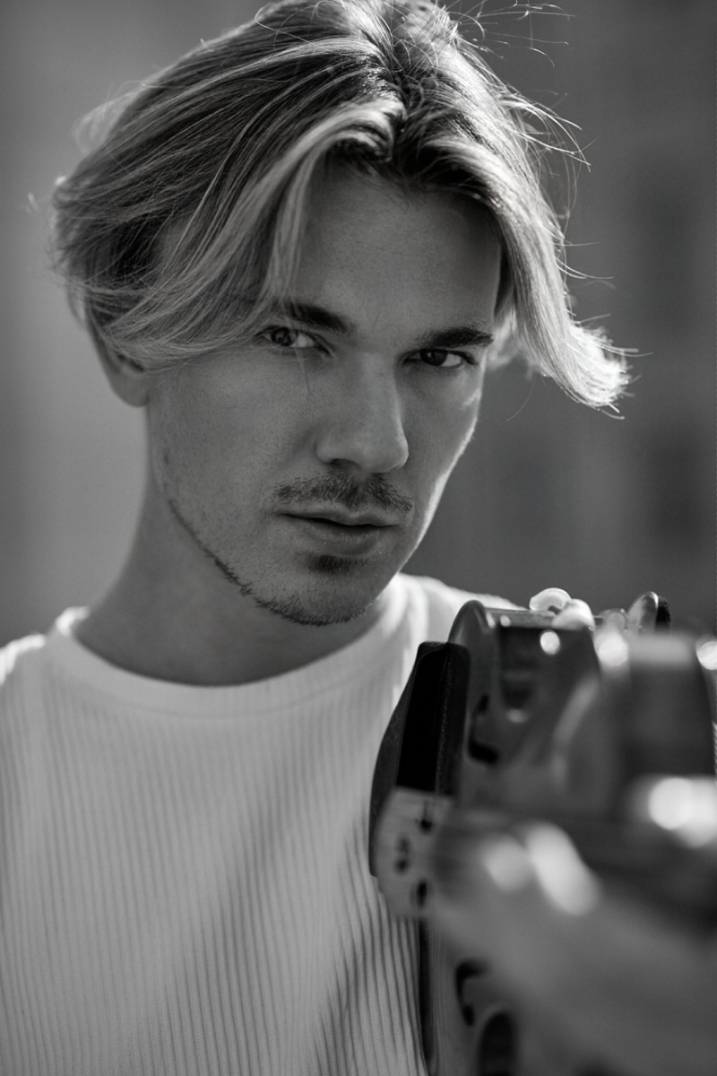 Junger Mann mit blonden, langen Haaren, tr&auml;gt ein wei&szlig;es Shirt und h&auml;lt ein Instrument in der Hand, Blick direkt in die Kamera.