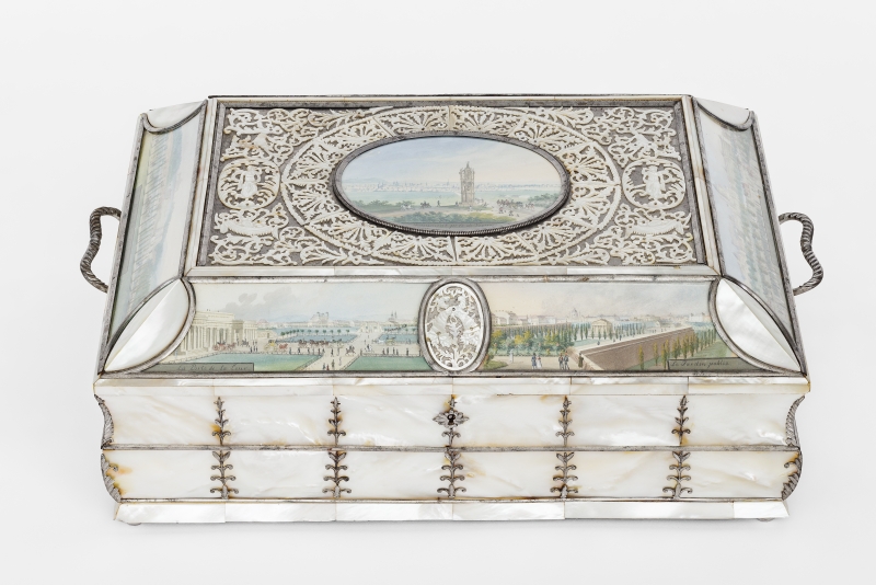 This box was made around the year 1825.
The watercolors on the box were painted by Balthasar Wigand.
The box is made of mother-of-pearl and silver.
It looks elegant and valuable.
On the sides, it has small handles for carrying.
On the lid, there is a small picture in the center:
It shows the Spinnerin am Kreuz – a well-known landmark in Vienna.
In the background, you can see the city of Vienna and people on horseback.
Around the picture, there is a fine metal decoration with leaves and small figures.
Painted scenes of Vienna also appear on the sides of the box –
specifically the outer city gate (Burgtor) and Heldenplatz.