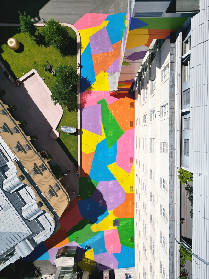 The photo shows the Hanuschhof from above. The floor is painted in bright colors - you can see large areas in blue, yellow, green, pink, orange and purple.

Many small, colorful dots can be seen in each colored area. These dots show how much CO₂ was emitted in a given year - one dot represents one billion tons.

The colors are vibrant and glow in the sunlight. The painting makes the courtyard friendly, eye-catching and lively - and it has a purpose:
The special paint helps to reduce the heat because it reflects the sunlight better than normal concrete.

This work of art is called “CO₂ntext”. It combines environmental issues, art and urban space - and invites people to reflect and linger.