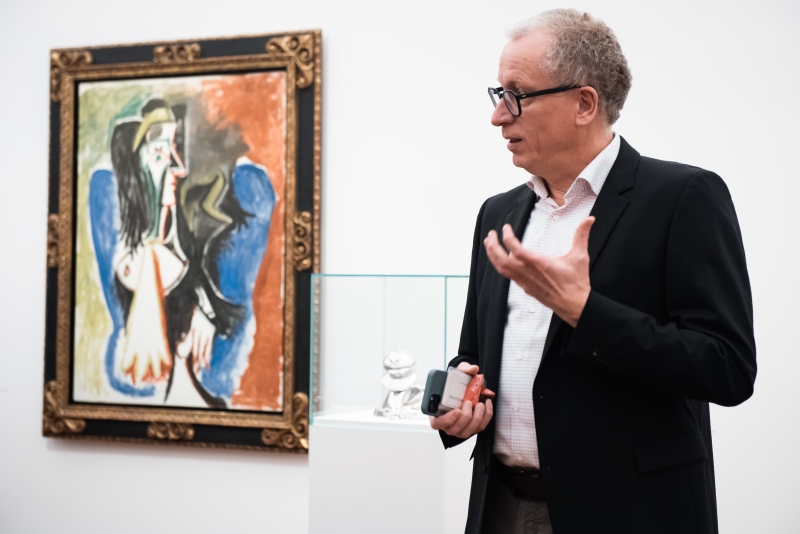Mann in Anzug spricht vor einem Picasso-Gem&auml;lde und einer Glasvitrine mit einer Skulptur.