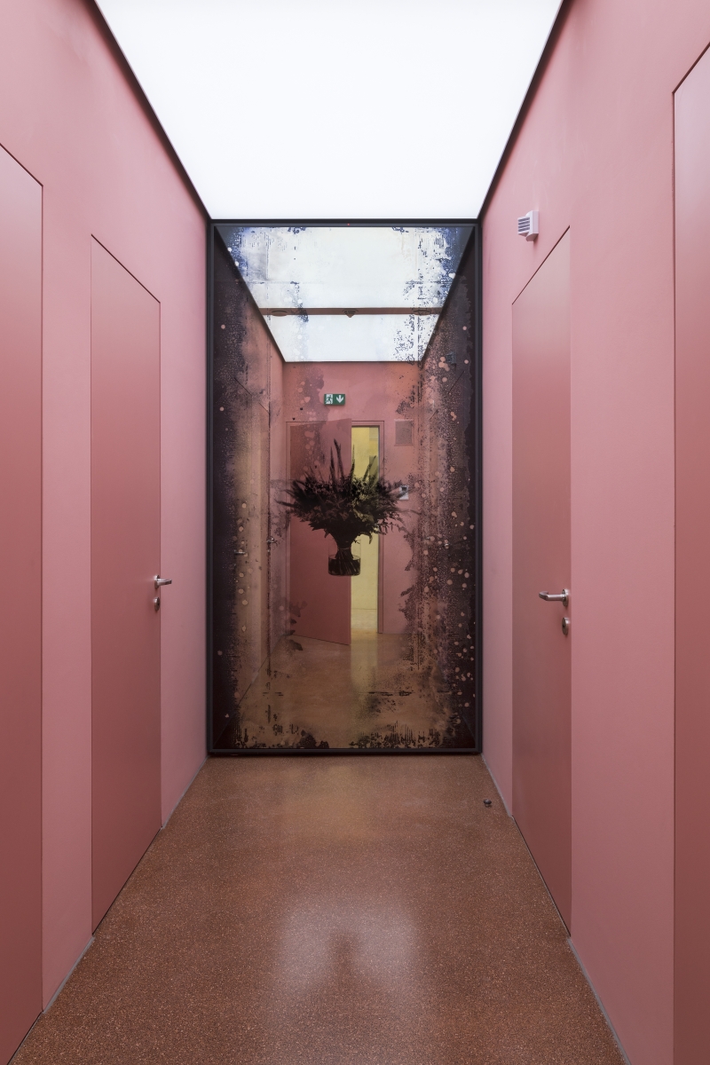 Auf dem Bild sehen wir einen Raum mit einem Spiegel mit einem Blumenstrauß in der Mitte.
Der Raum hat links und rechts rosa gestrichene Wände und Türen.
Der Spiegel ist am Ende des Raumes. Die Ränder des Spiegel sind dunkel und gesprenkelt, wie bei einem alten Foto oder Film.
Das Kunstwerk ist von Andreas Duscha.
Er hat den Spiegel selbst hergestellt, nach einem alten Rezept aus dem 17. Jahrhundert.
In der Mitte sieht man ein Blumenarrangement – es ist ein Siebdruck auf der Spiegelfläche.
Die Blumen auf dem Spiegel haben eine tiefere Bedeutung:
Sie stehen für gesellschaftliche Krisen und Revolutionen.
Ein Strauß erinnert zum Beispiel an Revolutionen: zum Beispiel für die Nelkenrevolution in Portugal oder
die Tulpenrevolution in der Ukraine.
Duscha interessiert sich für versteckte Geschichten und Zusammenhänge.
Er nutzt alte Techniken und besondere Materialien,
um Themen wie Macht, Wandel und Geschichte auf eine leise, poetische Weise sichtbar zu machen.
Dieses Kunstwerk gehört zu einer Serie von vier Spiegeln,
die im Vorraum der Toiletten des Museums der Heidi Horten Collection hängen.
Jeder Spiegel erzählt eine eigene Geschichte – durch Pflanzen und Symbole.