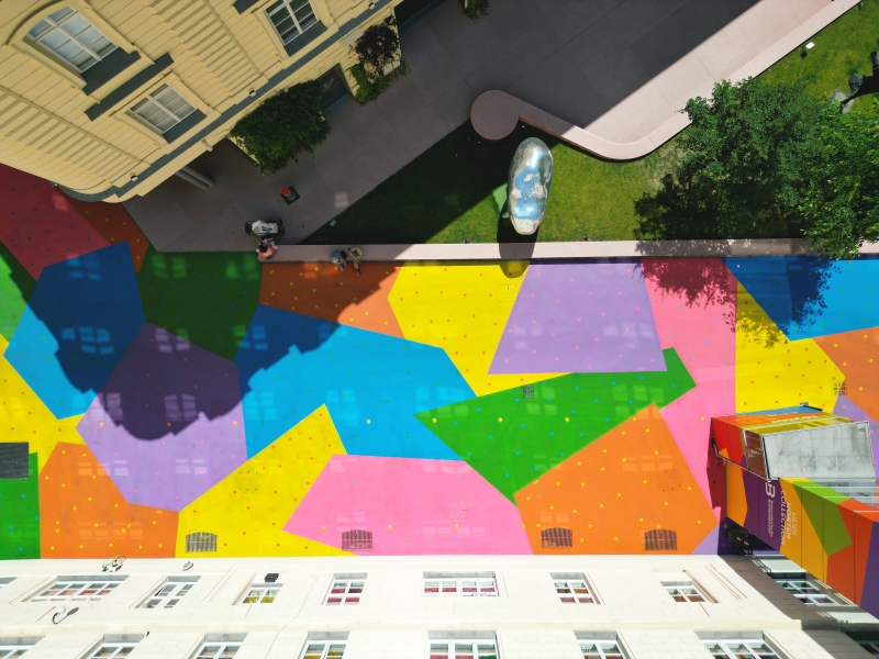The photo shows the Hanuschhof in Vienna -
where the Heidi Horten Collection museum is located.
The floor between the buildings is brightly painted.
You can see many colored areas in pink, blue, yellow, green, orange and purple.
These colors are not only beautiful, they also have an important meaning.
The painting shows how CO₂ emissions increase each year.
Each colorful field represents a year - the further you go, the more CO₂.
The color on the ground is also special:
It reflects sunlight better than dark asphalt.
This makes the square less hot - which helps the climate in the city.
On the top right of the picture you can see a small green park with works of art
and the bright museum building on the right.