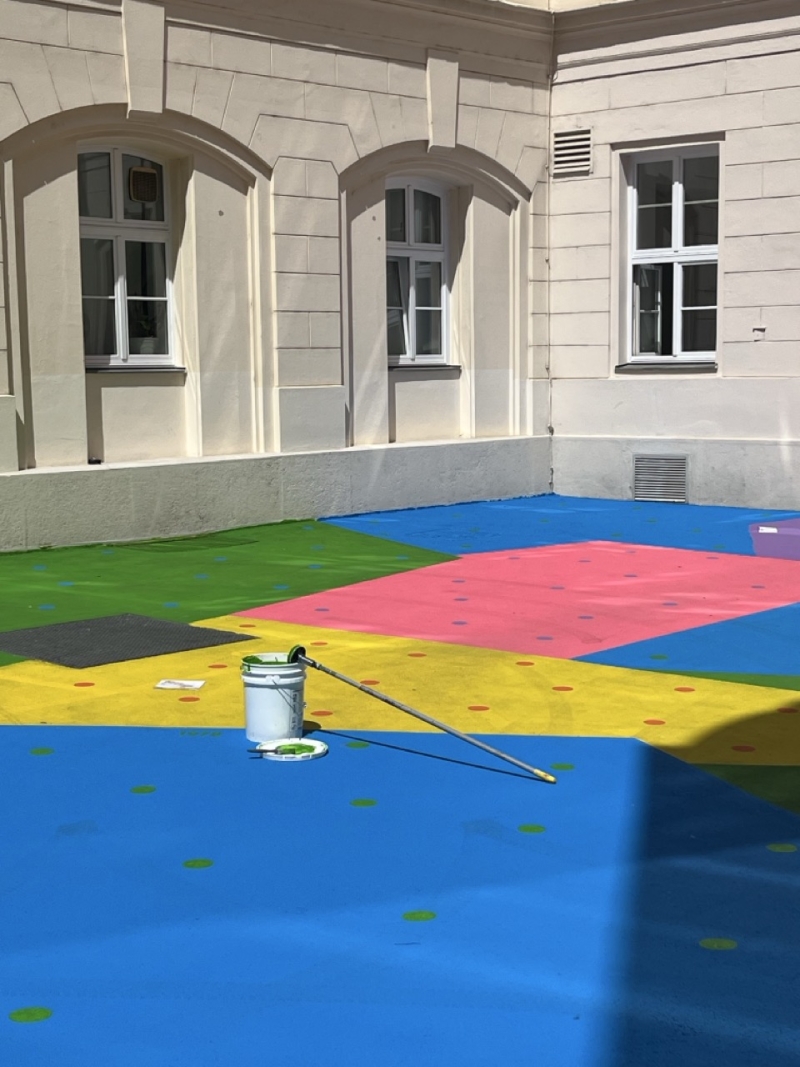 The photo shows the Hanuschhof in Vienna. The floor is painted in bright colors - you can see large areas in blue, yellow, green, pink and purple.
Many small, colorful dots can be seen in each colored area. These dots show how much CO₂ was emitted in a given year - one dot represents one billion tons.
At the front in the middle is a white paint bucket with a long handle, next to it is a small paint tray - you can see that the artwork is currently being worked on or has recently been completed.
The colors are vibrant and glow in the sunlight. The painting makes the courtyard friendly, eye-catching and lively - and it has a purpose:
The special paint helps to reduce the heat because it reflects the sunlight better than normal concrete.
This work of art is called “CO₂ntext”. It combines environmental issues, art and urban space - and invites people to reflect and linger.