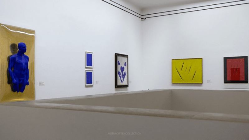 Blaue Skulptur an Wand, umgeben von farbigen Gemälden in einem modernen Ausstellungsraum.
