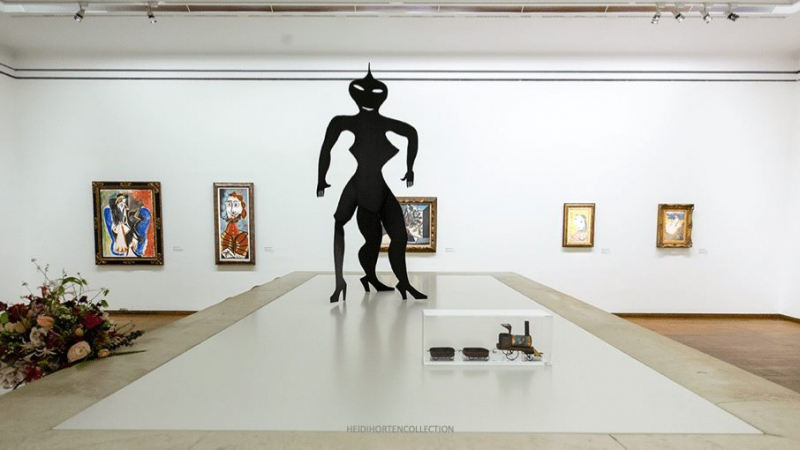 Schwarze Skulptur in der Mitte, umgeben von Gemälden an der Wand und einer Vitrine mit Kunstobjekten.