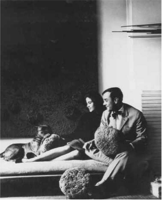 A man and woman sit on a couch, holding textured sculptures, with a dog beside them, against a dark abstract background.