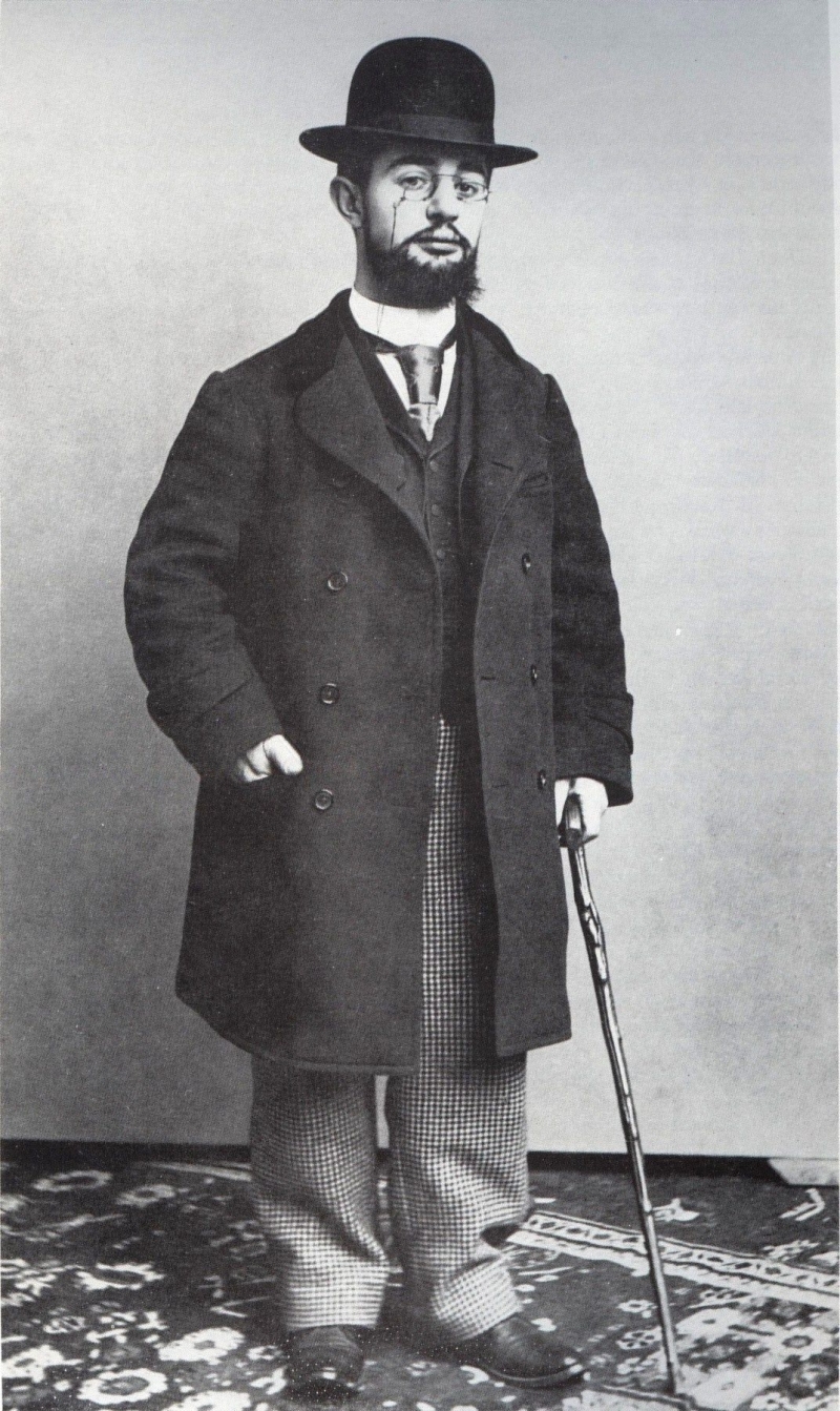 Das Foto zeigt den Künstler Henri de Toulouse-Lautrec. Er steht auf einem gemusterten Teppich vor einer schlichten Wand. Toulouse-Lautrec trägt einen dunklen Mantel, eine kariert gemusterte Hose und einen Hut. Er hat einen Schnurrbart und Bart und blickt ruhig in die Kamera. In seiner rechten Hand hält er einen Spazierstock, seine linke Hand steckt locker in der Manteltasche. Henri de Toulouse-Lautrec war ein französischer Maler und Plakatkünstler und ist bekannt für seine Darstellungen des Pariser Nachtlebens im 19. Jahrhundert. Er war kleinwüchsig und hatte durch eine Erkrankung Schwierigkeiten beim Gehen – darum nutzte er oft einen Stock zur Unterstützung.