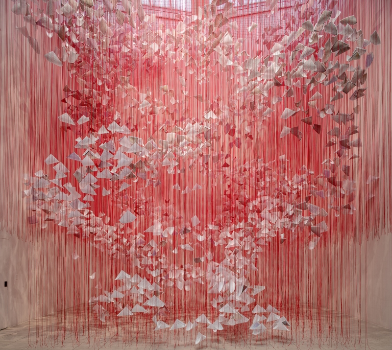 The installation fills an exhibition space with a dense web of bright red threads stretching from the ceiling to the floor. Hundreds of white sheets of paper float within this net - they look like folded letters, loose sheets or small wings. The letters spiral upwards or outwards as if in a movement of air. The red color of the threads dominates the room. The threads are so numerous and dense that from a distance they look like a shimmering curtain that changes the boundaries of the room and creates a walk-in, almost physical experience.