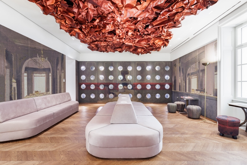 The photo shows the Tea Room in the Heidi Horten Collection museum in Vienna.
A large, red work of art by Hans Kupelwieser hangs from the ceiling.
It looks like crumpled paper or shiny fabric, but
is made of aluminum.
There are lots of round windows on the back wall - they look like portholes.
Behind them are small, precious objects from Heidi Horten's collection.
This showcase wall was designed by Markus Schinwald.
The pink seating in the middle of the room is also part of his concept.
They invite you to sit down, look and relax.
The walls are covered with large fabric pictures,
reminiscent of an old café or castle room.