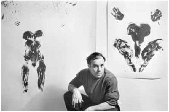 Yves Klein crouches beside two abstract black ink artworks on white walls, showcasing fluid, expressive forms.