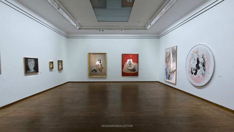 Zu sehen ist die Ausstellung "WOW" im Leopold Museum. In den Raum sieht man Werke von Georg Baselitz auf der rechten Seite. In der mittleren Wand sieht man Werke von Francis Bacon und auf der linken Wand sind Werke von Gerhard Richter.