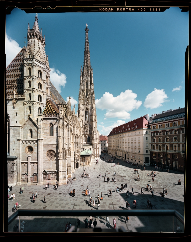 In the photo, we see St. Stephen’s Cathedral in Vienna.
The picture was taken from a balcony.
St. Stephen’s Cathedral is a very old church.
In the middle, you can see a very tall, pointed tower.
The roof has colorful tiles. It looks beautiful and special.
On the left is the church, on the right are other buildings.
There are many people on the square in front of it.
The sky is blue. There are a few white clouds.
The photo was taken with an old camera that uses film.
The photographer’s name is Stefan Oláh.
The colors in the picture are warm and pleasant.