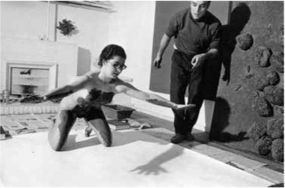 A woman with paint on her body reaches toward a large canvas, while a man observes her from the side in a studio setting.