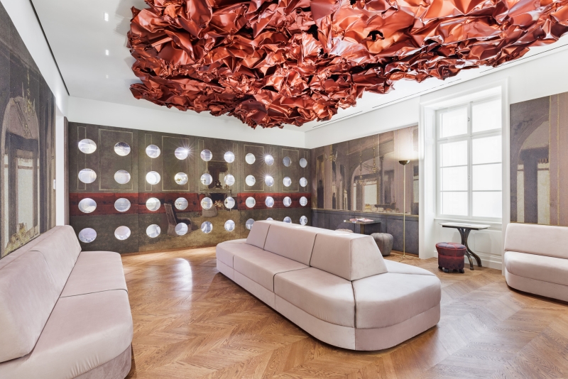 The photo shows the Tea Room in the Heidi Horten Collection museum in Vienna.

A large, red work of art by Hans Kupelwieser hangs from the ceiling.
It looks like crumpled paper or shiny fabric, but
is made of aluminum.

There are lots of round windows on the back wall - they look like portholes.
Behind them are small, precious objects from Heidi Horten's collection.

This showcase wall was designed by Markus Schinwald.
The pink seating in the middle of the room is also part of his concept.
They invite you to sit down, look and relax.

The walls are covered with large fabric pictures,
reminiscent of an old café or castle room.