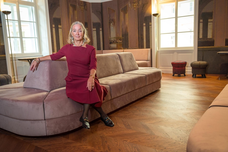 Agnes Husslein-Arco in rotem Kleid sitzt entspannt auf einer modernen, hellen Couch in einem eleganten Raum mit Holzfu&szlig;boden.