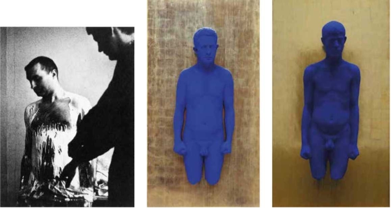 A black-and-white photo of a man with paint on his chest, flanked by two blue sculptures of a male figure.