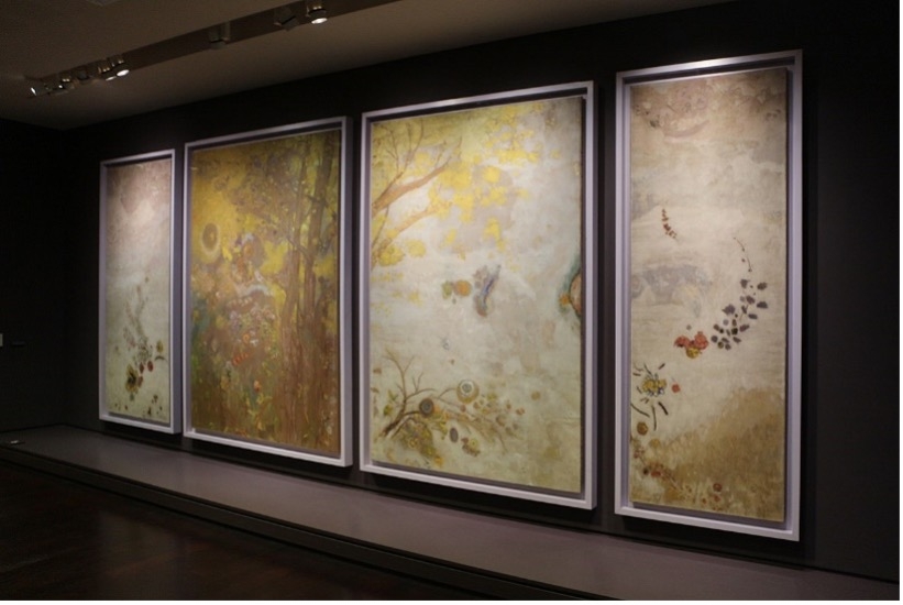 Vier gro&szlig;e, gerahmte Gem&auml;lde in sanften Farben, h&auml;ngen an einer dunklen Wand im Mus&eacute;e d'Orsay.
