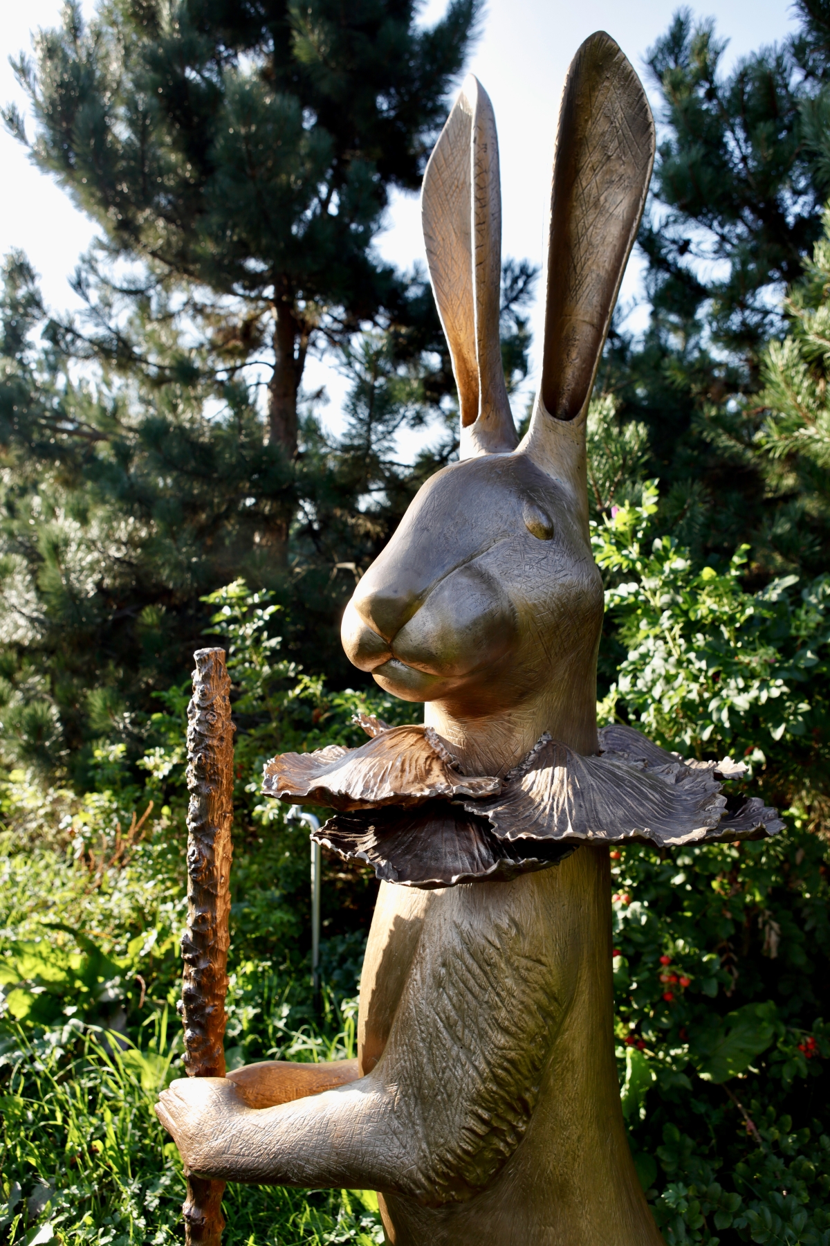 Bronzeskulptur eines Hasen mit einem Stock und einem runden Kragen aus Bl&auml;ttern, umgeben von gr&uuml;nen Pflanzen.