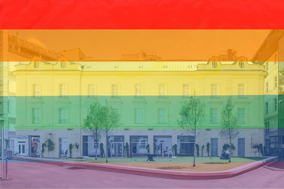 Fassade eines Geb&auml;udes mit Regenbogenfarben im Hintergrund, umgeben von B&auml;umen und einer kleinen Gr&uuml;nfl&auml;che.