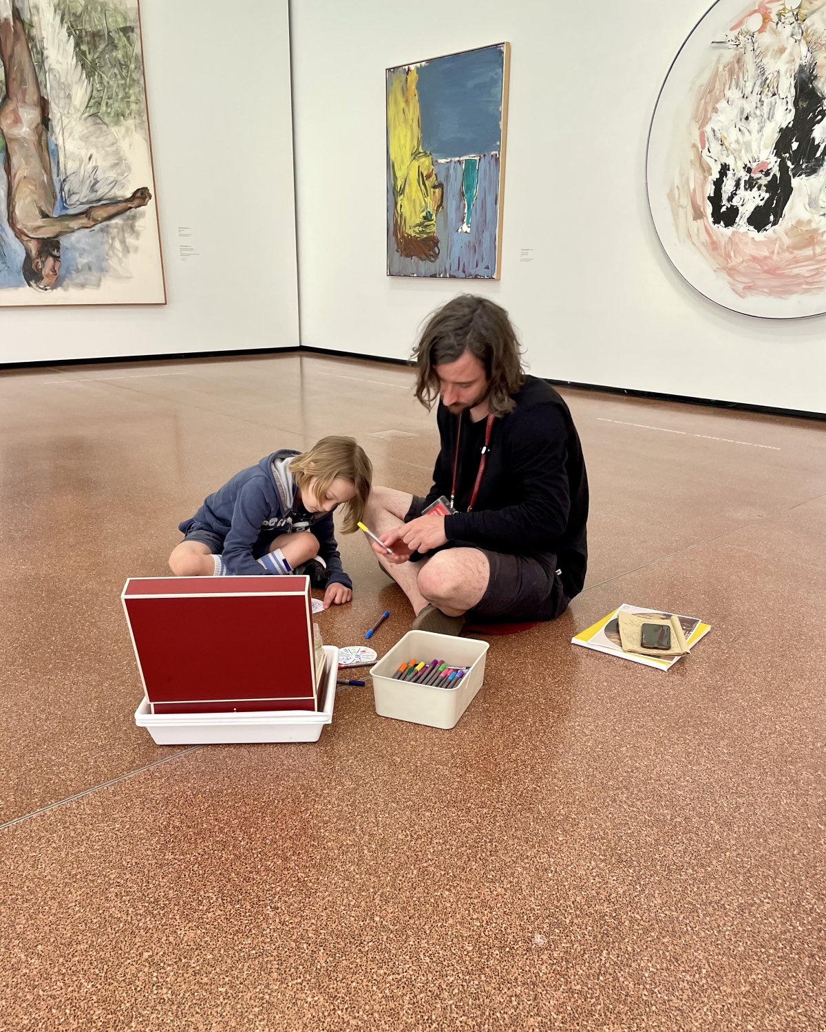 Ein Erwachsener und ein Kind sitzen auf dem Boden eines Kunstmuseums, umgeben von Gem&auml;lden, und zeichnen.