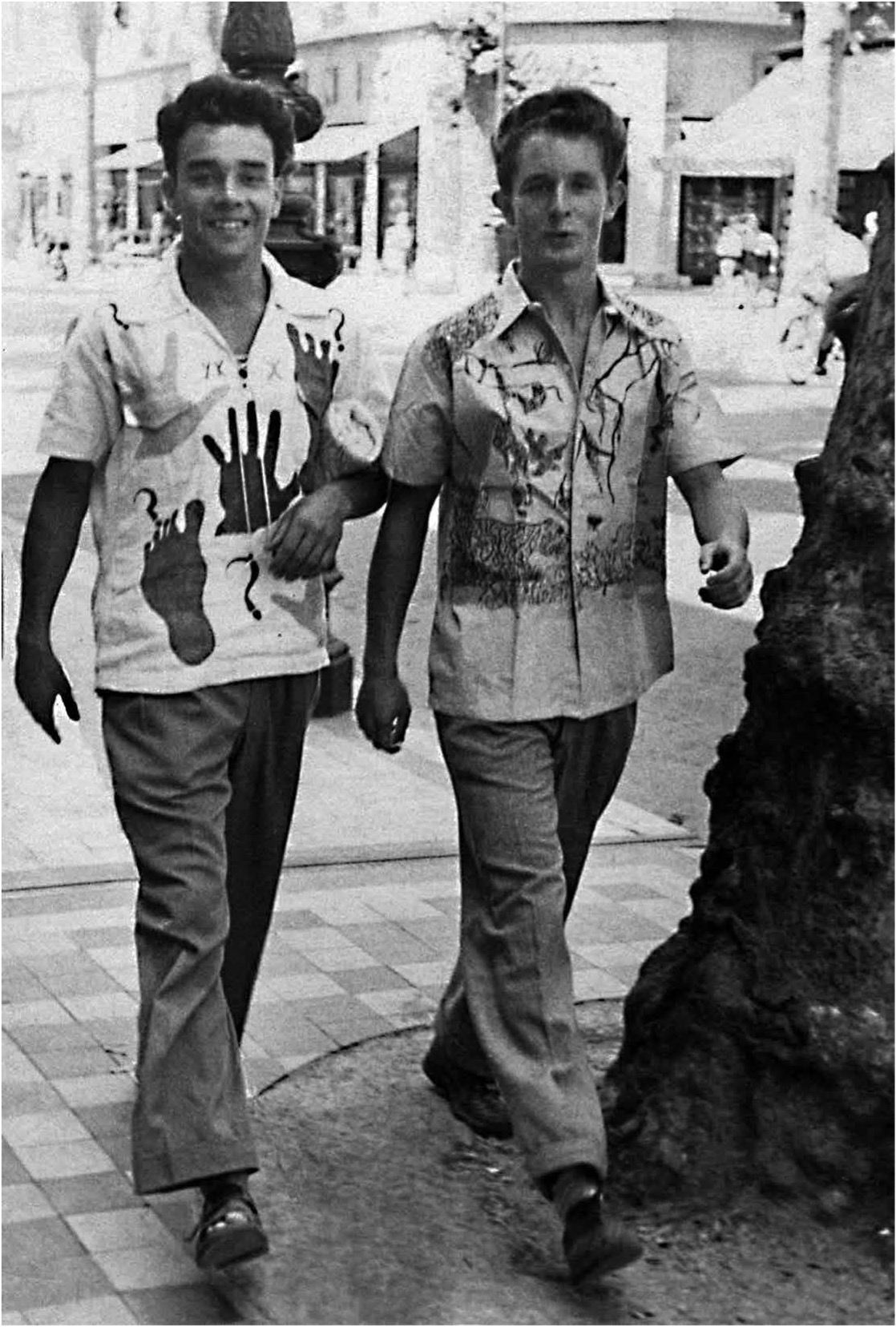 Two young men walk together on a city street, wearing colorful, patterned shirts and smiling at the camera.