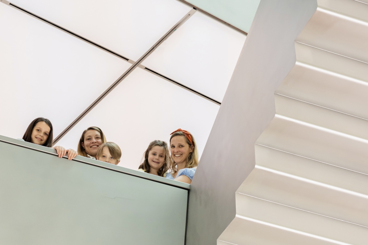 Auf dem Foto sieht man f&uuml;nf Personen, darunter drei Kinder und zwei Erwachsene. Sie stehen auf einer Ausstellungsebene und schauen freundlich in die Kamera. Alle l&auml;cheln.

Rechts im Bild sieht man eine moderne, helle Treppe. Hinter den Personen sieht man die wei&szlig;e Lichterdecke.