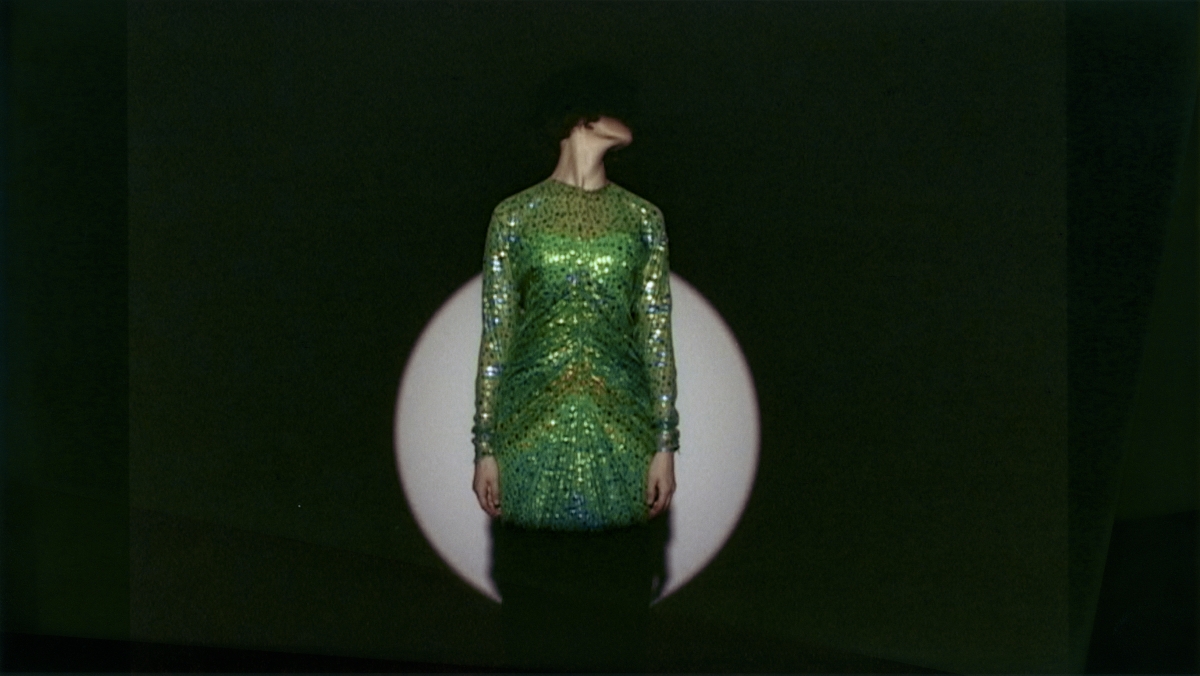 Auf dem Bild sieht man eine Person in einem gr&uuml;nen Kleid.
Das Kleid ist gl&auml;nzend und mit vielen Pailletten oder Glitzersteinen verziert.
Die Person steht vor einem dunklen Hintergrund,
in dem ein runder, heller Lichtkreis, wahrscheinlich von einem Scheinwerfer, zu sehen ist

Der Kopf der Person ist nach links geneigt,
sodass man das Gesicht nicht sehen kann.
Das macht das Bild geheimnisvoll und spannend.