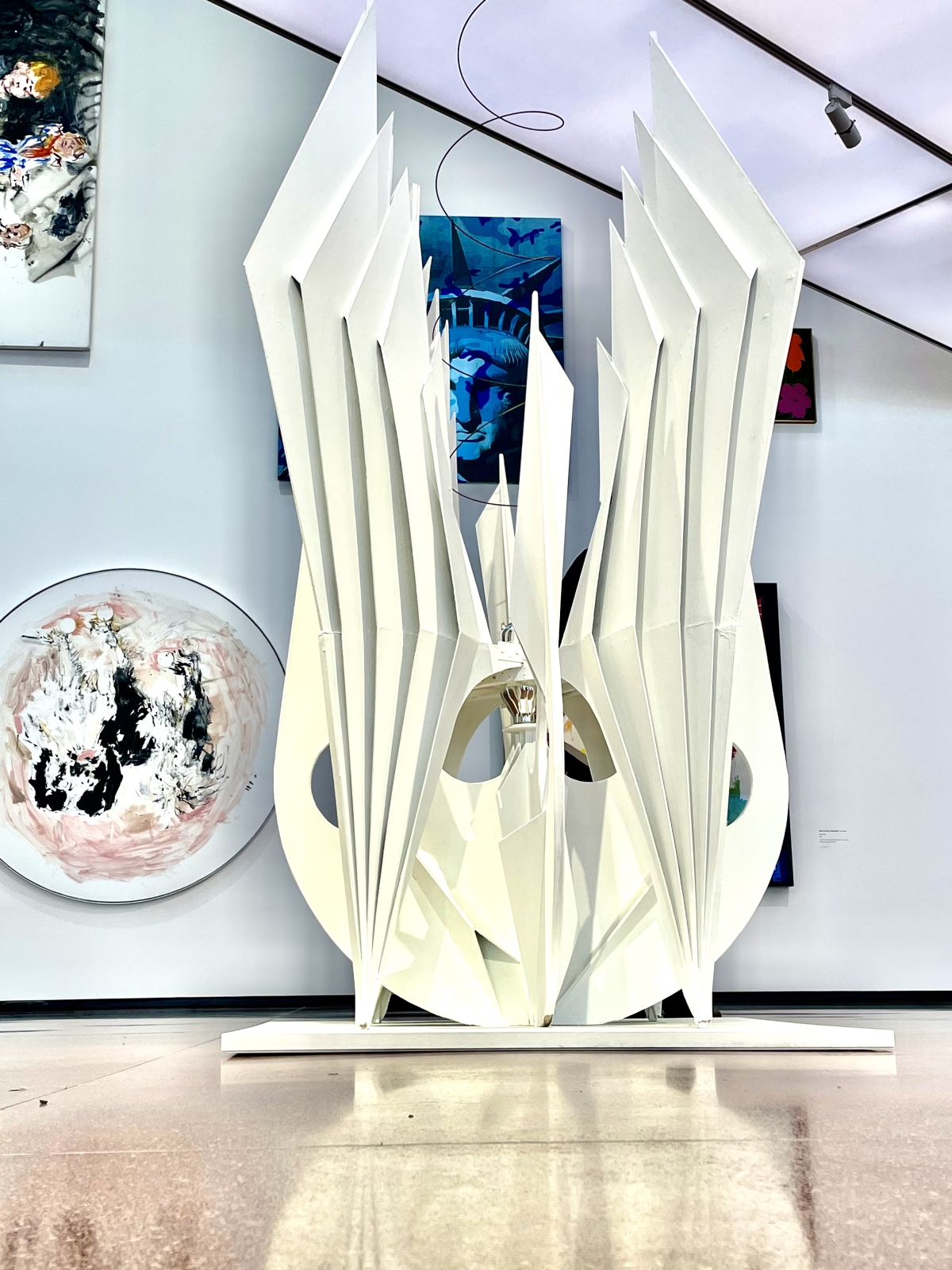 Skulptur mit wei&szlig;en, spitzen Elementen in einem modernen Ausstellungsraum, umgeben von bunten Gem&auml;lden.
